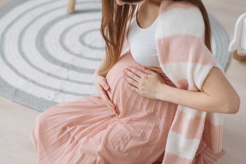 Pregnant woman sitting indoors, tenderly holding her baby bump in soft, cozy attire.