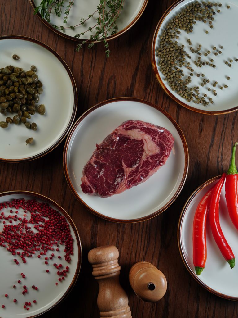 Appetizing flat lay of raw steak, peppers, spices, and herbs on a wooden table.