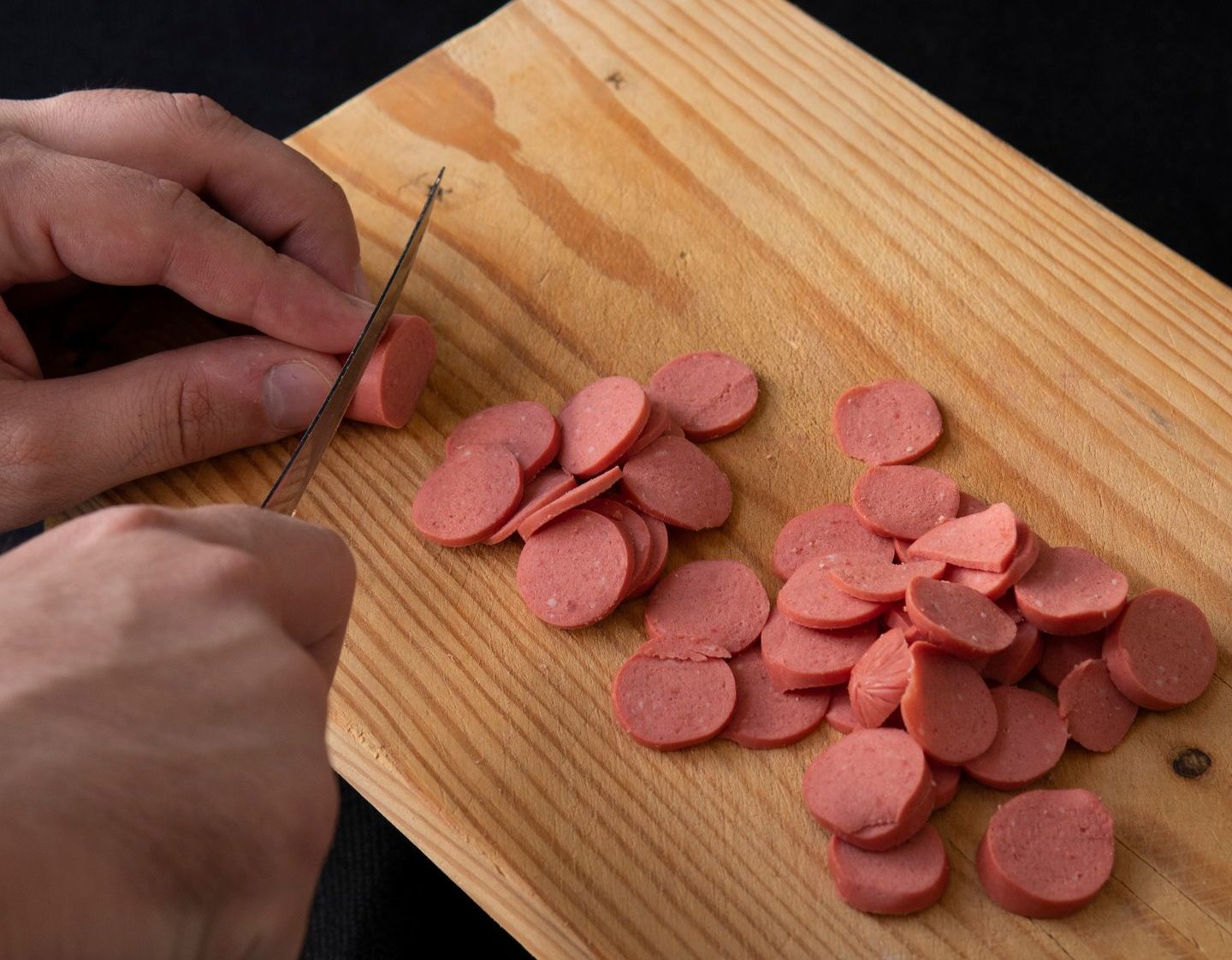 From above of crop unrecognizable cook cutting yummy sausage on wooden cutting board on black background