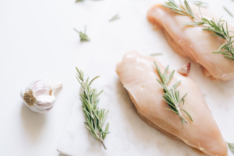 Raw chicken breasts garnished with rosemary and garlic on a white surface.